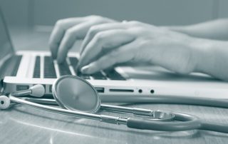 weibliche Hände die etwas an einem Laptop eintippen daneben liegt ein Stethoskop